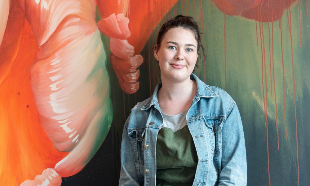 Emily, the author of the piece, stands in front of a mural smiling at the camera.