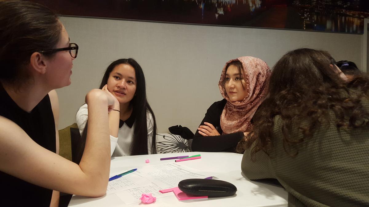 Students sit around a table having a conversation, there are pens and sheets of paper covered in written plans between them.