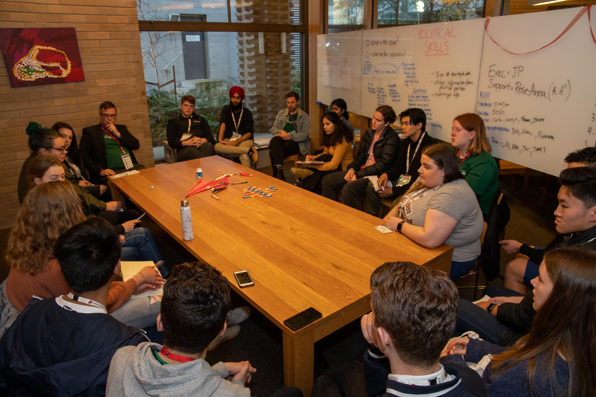 A group of students and facilitators sit around a table, a whiteboard in the background reads 'political skills'